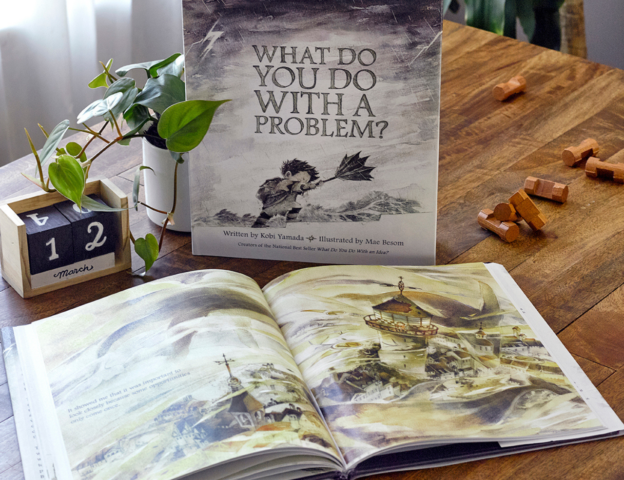 Compendium What Do You Do With a Problem book on a wooden table with an open copy and a plant