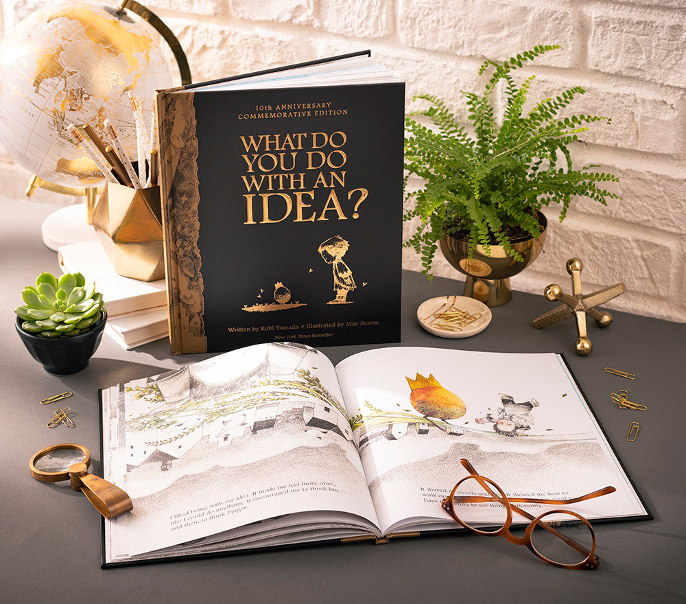 What Do You Do With an Idea? 10th Anniversary Edition book by Compendium, black and gold cover standing open on a desk with a fern and reading glasses