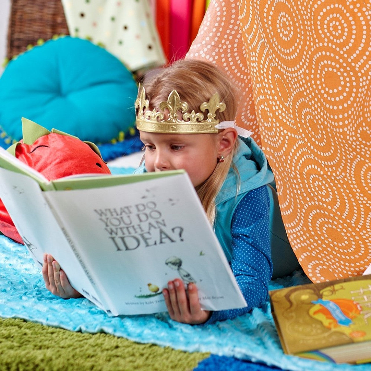 Girl in a paper crown reading Compendium What Do You Do With an Idea in a colorful blanket fort