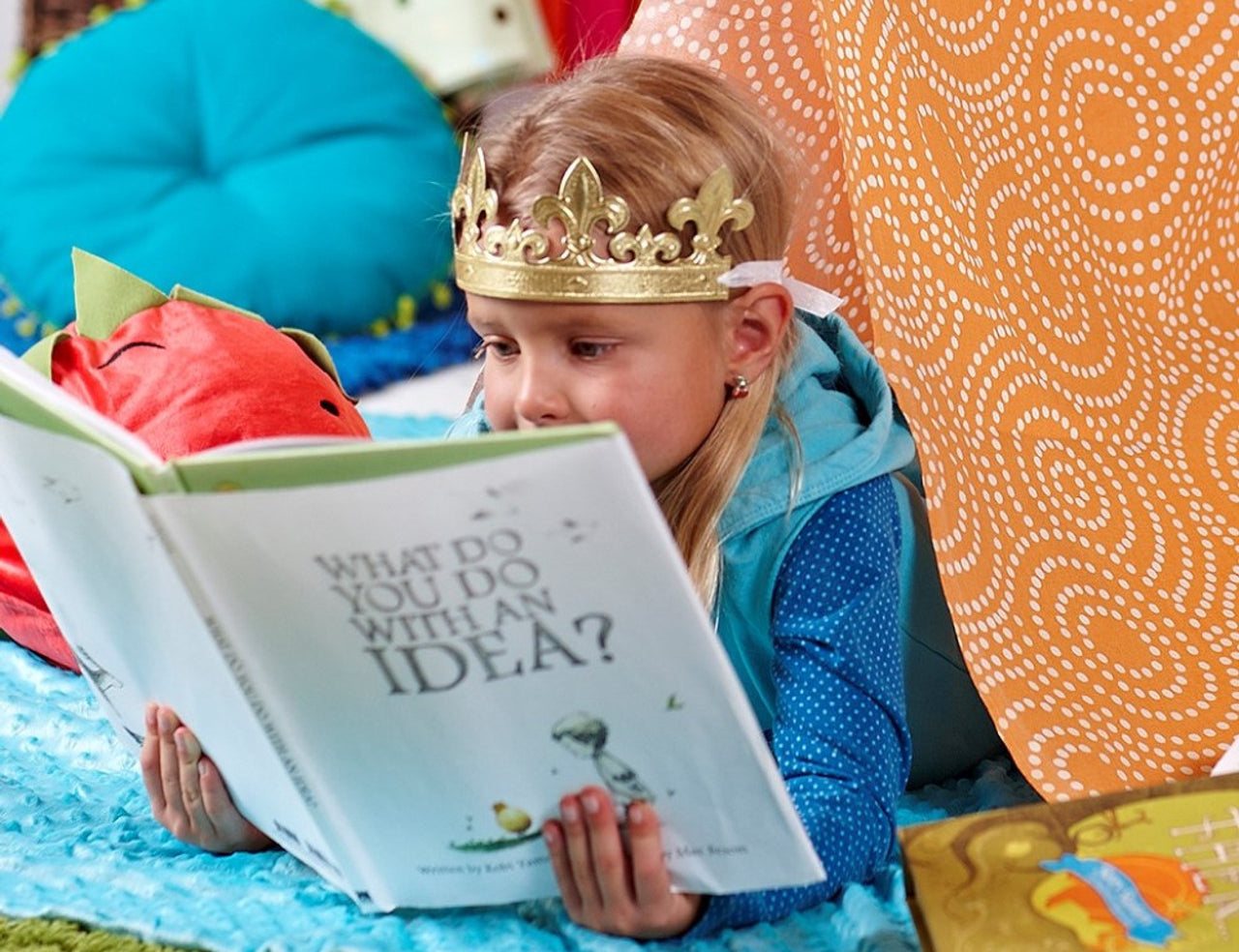Girl in a paper crown reading Compendium What Do You Do With an Idea in a colorful blanket fort