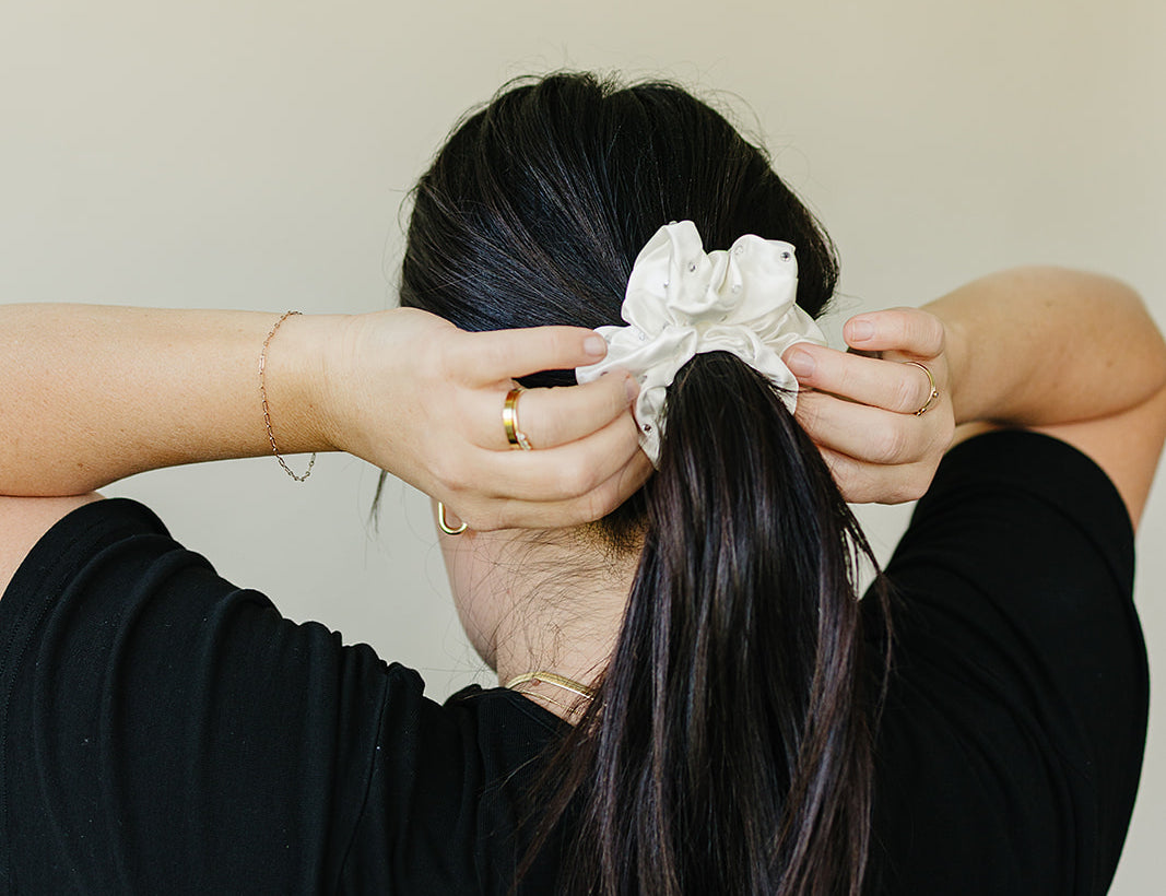 The Ivory crystal mulberry silk scrunchie, set with tiny rhinestones, tied around a long dark ponytail seen from behind.