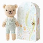 Cuddle + Kind Tiny Charlie the Honey Bear, a small hand-knit bear in mint overalls beside its floral arch box
