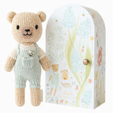 Cuddle + Kind Tiny Charlie the Honey Bear, a small hand-knit bear in mint overalls beside its floral arch box