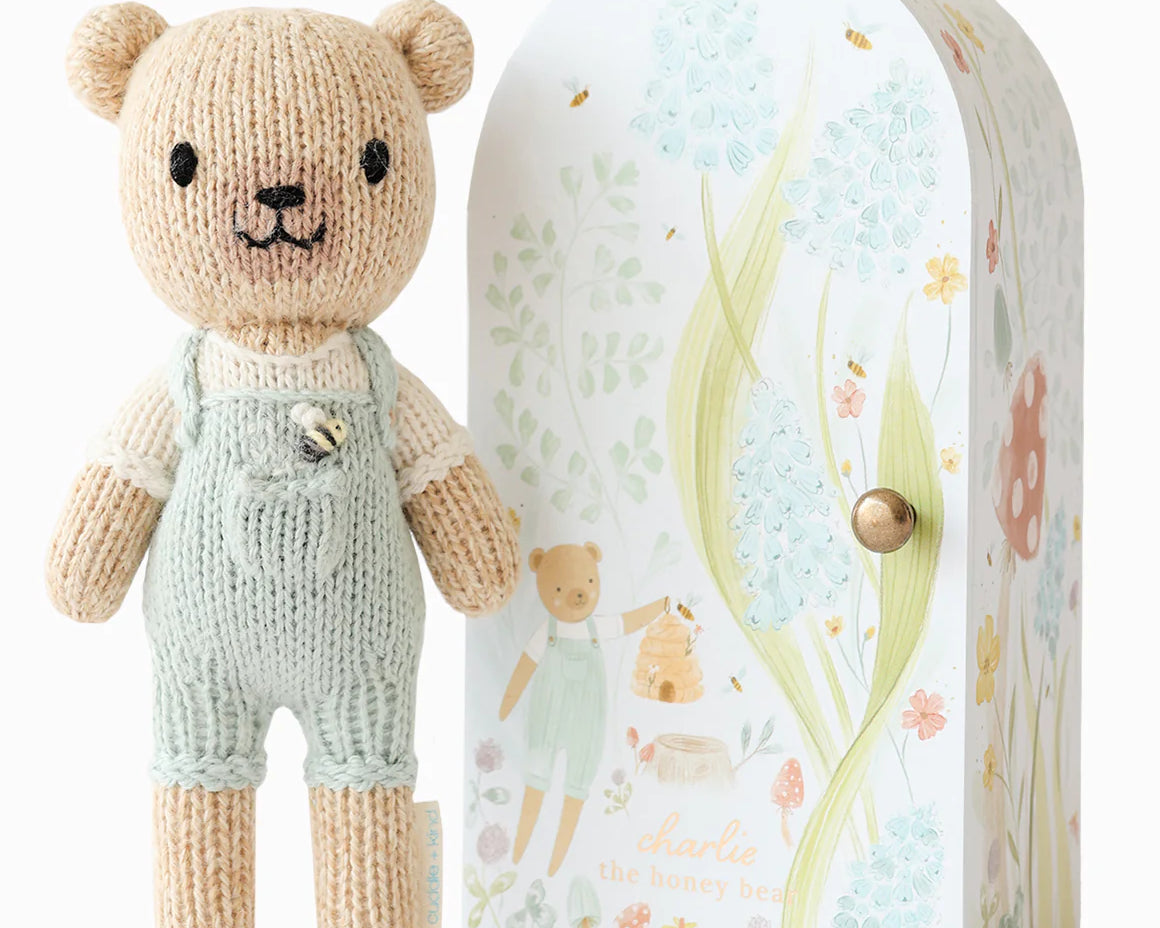 Cuddle + Kind Tiny Charlie the Honey Bear, a small hand-knit bear in mint overalls beside its floral arch box