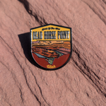 Shield shaped Dead Horse Point enamel pin showing the Colorado River gooseneck below the overlook, lying on red sandstone in Moab, Utah.