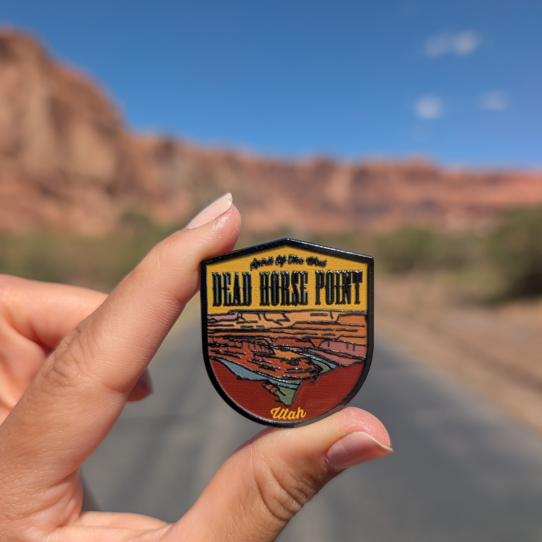 Hand holding the Dead Horse Point enamel pin on a road through the Moab red rock cliffs.