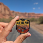 Hand holding the Dead Horse Point enamel pin on a road through the Moab red rock cliffs.