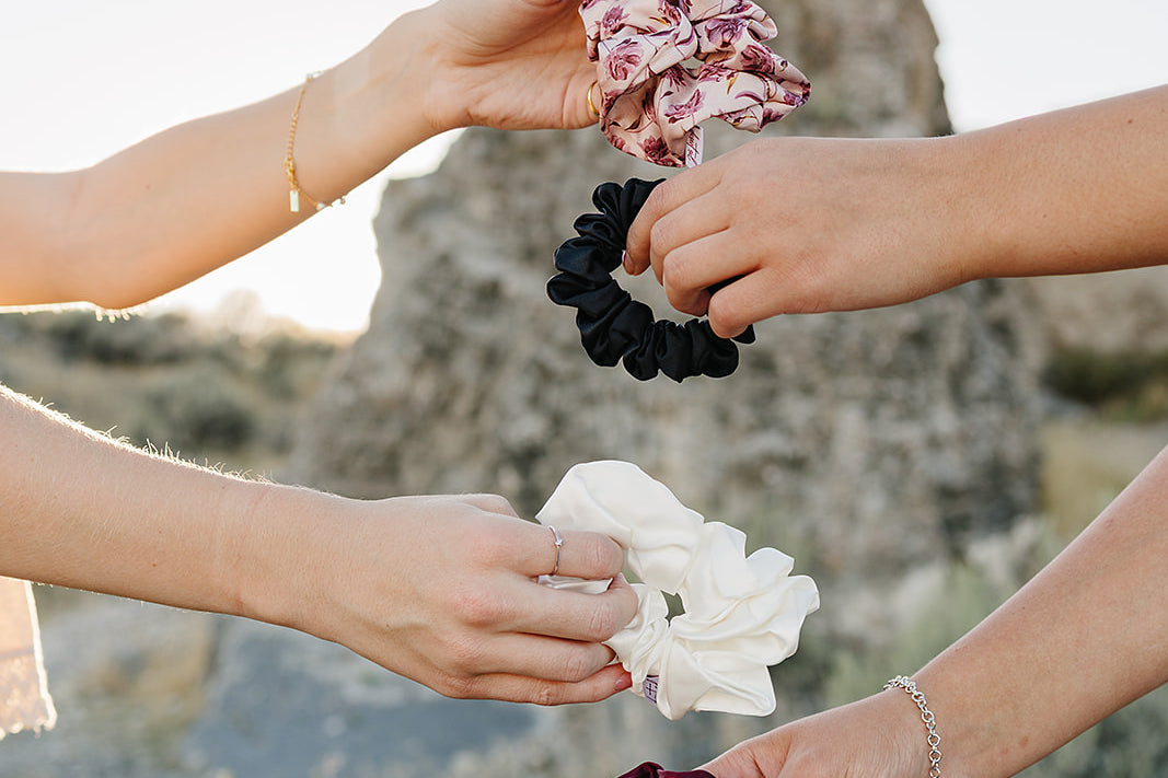 Four hands holding Desert Sol scrunchies in Mulberry Floral, black, ivory and plum crystal against a desert butte