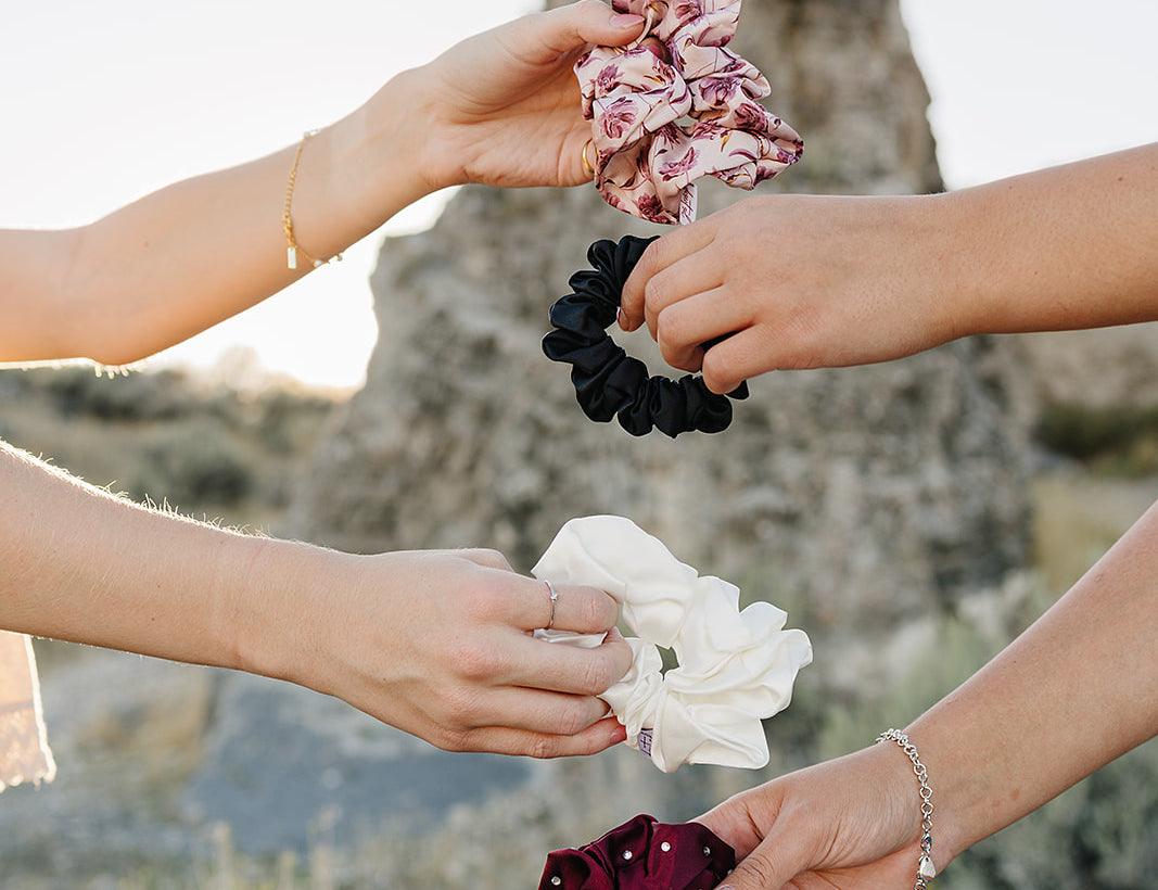 Four hands holding Desert Sol scrunchies in Mulberry Floral, black, ivory and plum crystal against a desert butte