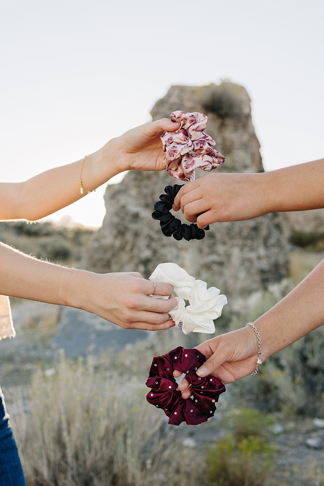 Four hands holding Desert Sol scrunchies in Mulberry Floral, black, ivory and plum crystal against a desert butte