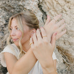 Model with stacked gold rings including the Desert Sol Meadow Ring and a Solara pearl bracelet, hands raised against desert rock
