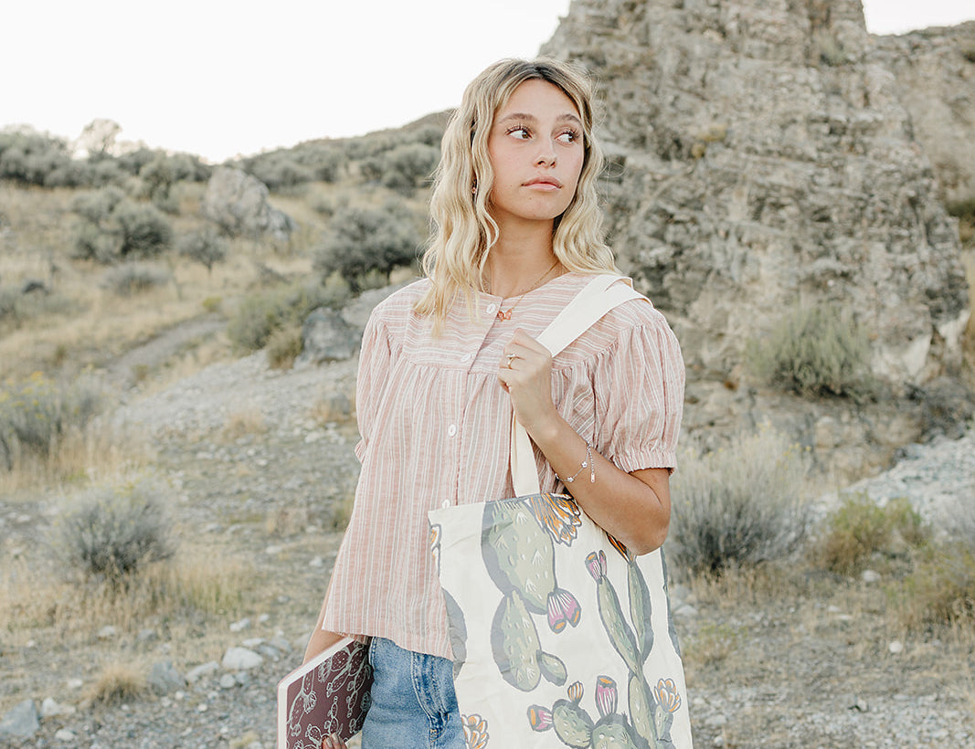 Model in a pink striped top carrying the Desert Sol Carryall Tote in Cactus Bloom with a plum journal in hand in the desert