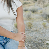 Desert Sol Wildflower Daisy Chain Bracelet and Wild Bloom Bracelet in gold on a model kneeling in the desert