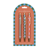 A Dopo Pen Set of 3, three patterned ballpoint pens, one gold, one green striped and one pink patterned, on a blue and green checkered backer card