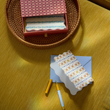 Designworks Notecards Set of 16, red patterned box in a woven basket with a scalloped card, blue envelope and pens on a yellow cloth