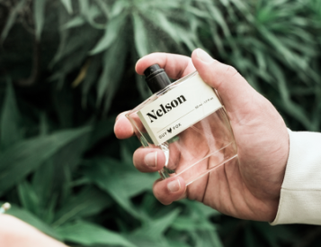 Hand holding the Guy Fox Nelson Men's Cologne bottle among green foliage