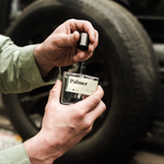 Hands uncapping the Guy Fox Palmer Men's Cologne bottle beside a truck tire