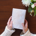Hands holding the Hosanna Revival NLT Compact Bible Casablanca theme over a wooden table with flowers