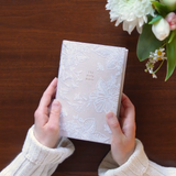Hands holding the Hosanna Revival NLT Compact Bible Casablanca theme over a wooden table with flowers