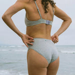 Back view of a woman at the beach wearing the Huha Mineral Bikini underwear and bralette in Grey