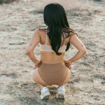 Woman with long dark hair crouching on sand, back view, wearing the Huha Mineral Bikini underwear in Tan