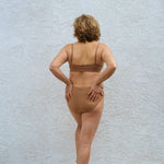Back view of a woman against a white wall wearing the Huha Mineral Bikini underwear in Tan