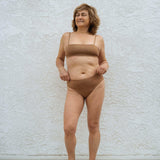 Woman standing against a white wall wearing the Huha Mineral Bikini underwear and bralette in Tan