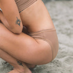 Close-up of a woman crouching on rocks wearing the Huha Mineral Bikini underwear in Tan