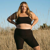 Woman standing in a field wearing the Huha Tata Bralette and bike shorts in Black, front view