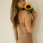 Woman holding a sunflower over her shoulder wearing the Huha Tata Bralette in Tan, back view