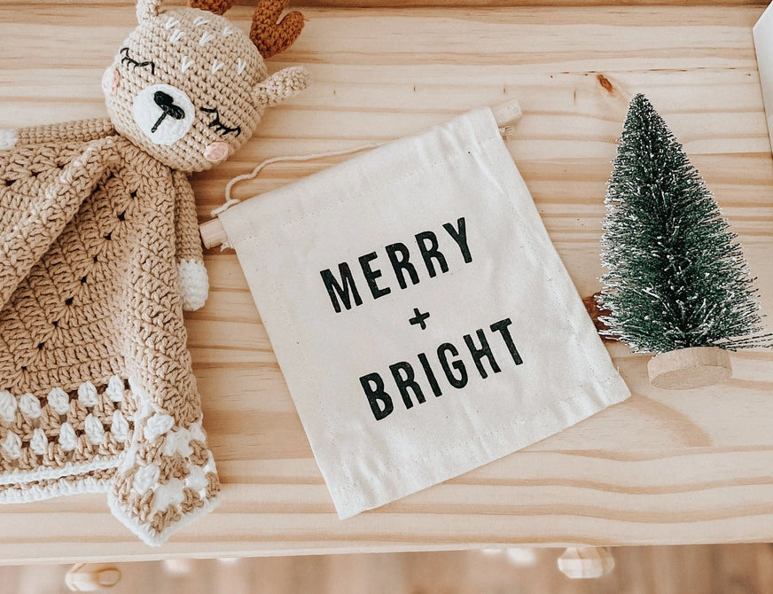 Merry + Bright Hanging Sign, second view, the canvas banner on a wood table with a bottle brush tree and a knit deer lovey