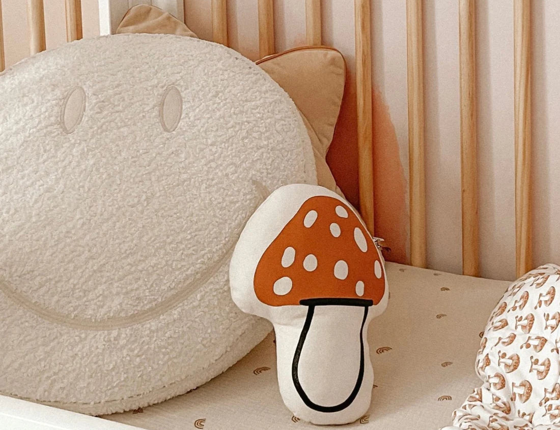 Imani Collective Mushroom Pillow in a crib beside a smiley face pillow