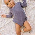 Baby in a purple onesie lying on the Jujube Roarsome Swaddle Blanket with a dinosaur print