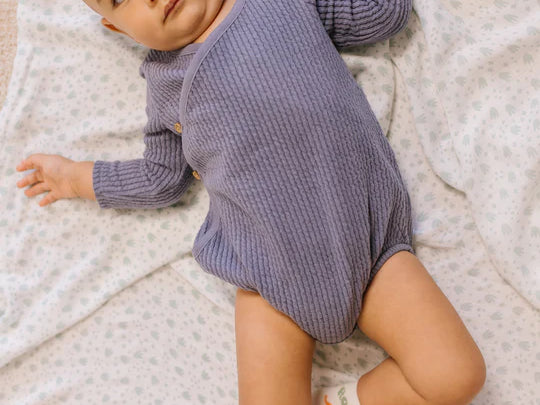 Baby in a purple onesie lying on the Jujube Roarsome Swaddle Blanket with a dinosaur print
