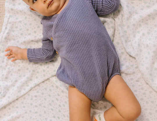 Baby in a purple onesie lying on the Jujube Roarsome Swaddle Blanket with a dinosaur print