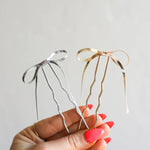 Hand holding two Kaxi Bow Pins, one silver and one gold, thin metal bow-shaped hair pins