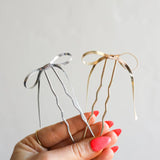 Hand holding two Kaxi Bow Pins, one silver and one gold, thin metal bow-shaped hair pins