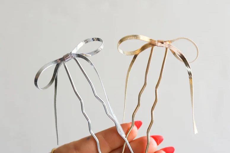 Hand holding two Kaxi Bow Pins, one silver and one gold, thin metal bow-shaped hair pins