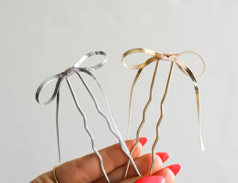 Hand holding two Kaxi Bow Pins, one silver and one gold, thin metal bow-shaped hair pins
