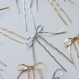 Several Kaxi Bow Pins in silver and gold scattered on a white surface