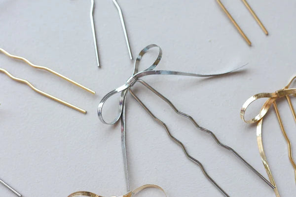 Several Kaxi Bow Pins in silver and gold scattered on a white surface
