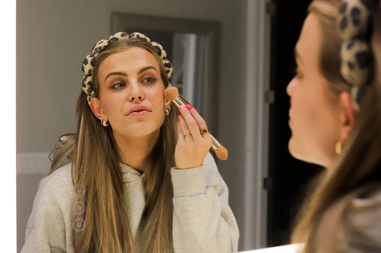 Woman applying makeup in a mirror wearing the Kaxi GRWM Headband in Cheetah