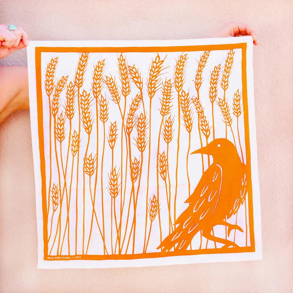 Kei & Molly Cotton Bandana in Raven, an orange bandana with a raven in a wheat field print, held flat