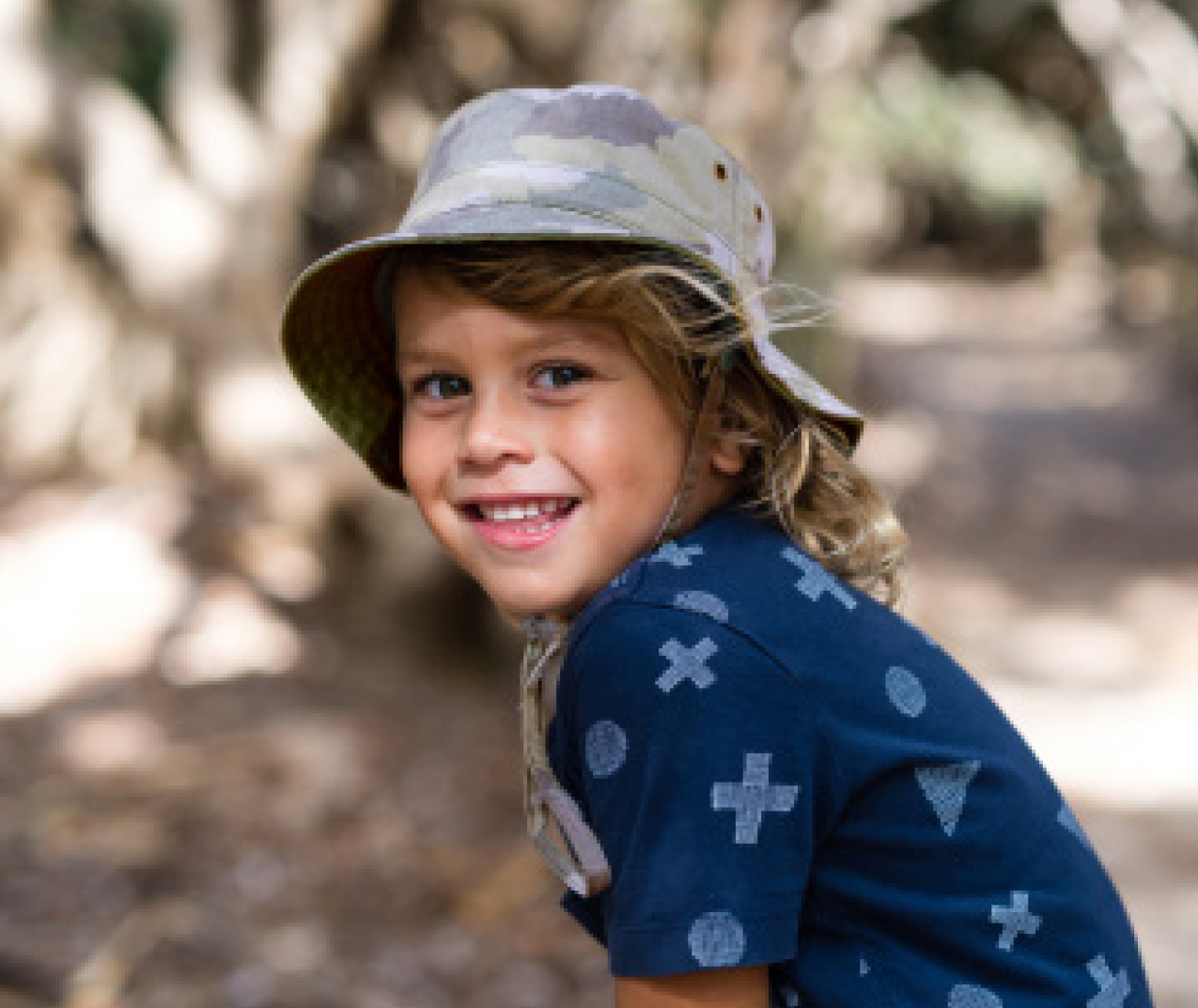 Boy wearing the Kooringal Dozer Boys Bucket Hat in Camo outdoors under trees