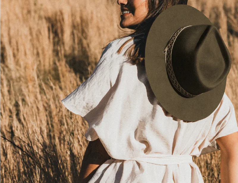 Woman in a linen dress in a wheat field wearing the Kooringal Women's Wide Brim Phoenix hat in Olive, looking back