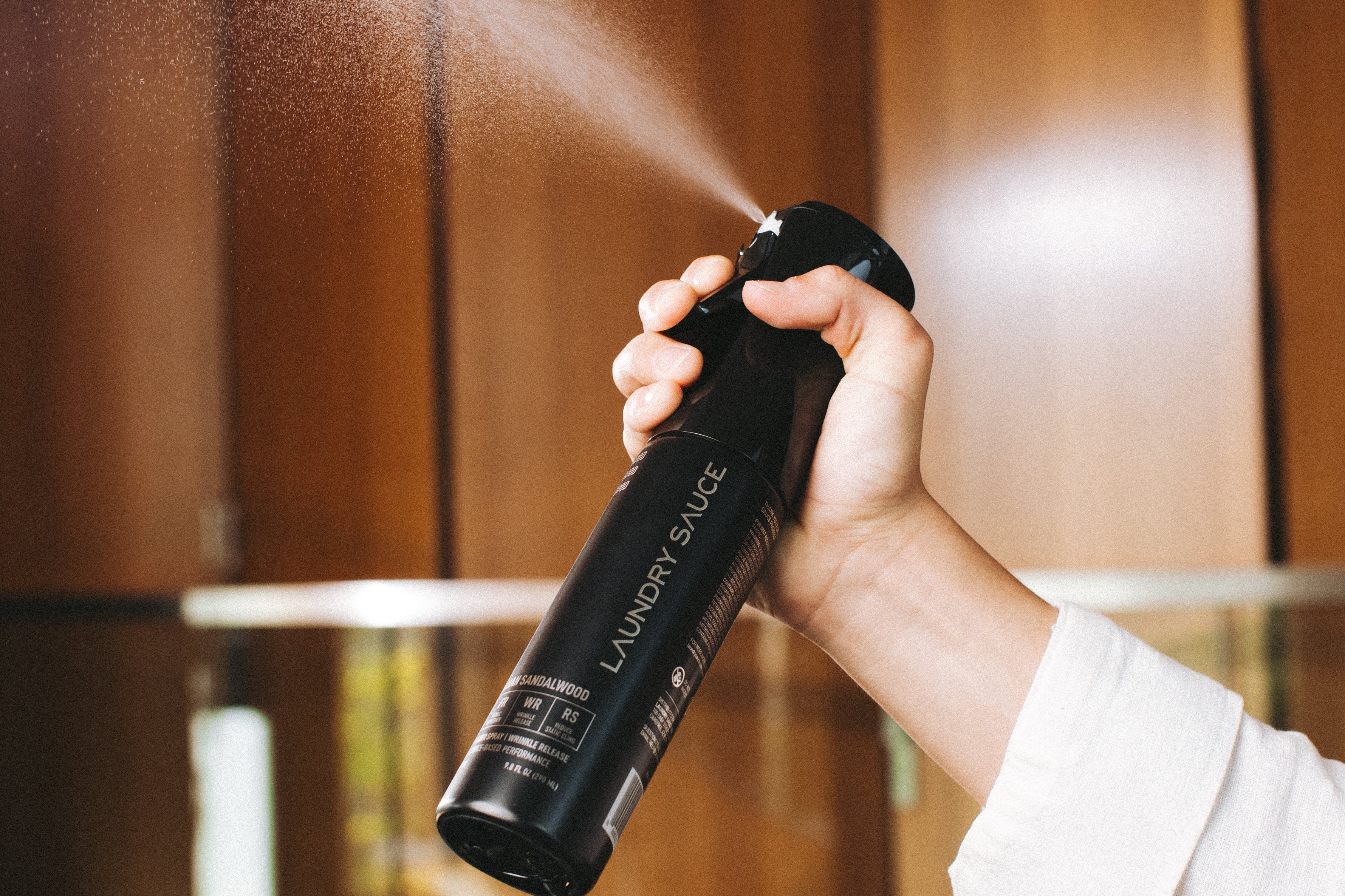 Hand spraying the Laundry Sauce Fabric Refresher Spray in a wood-paneled closet