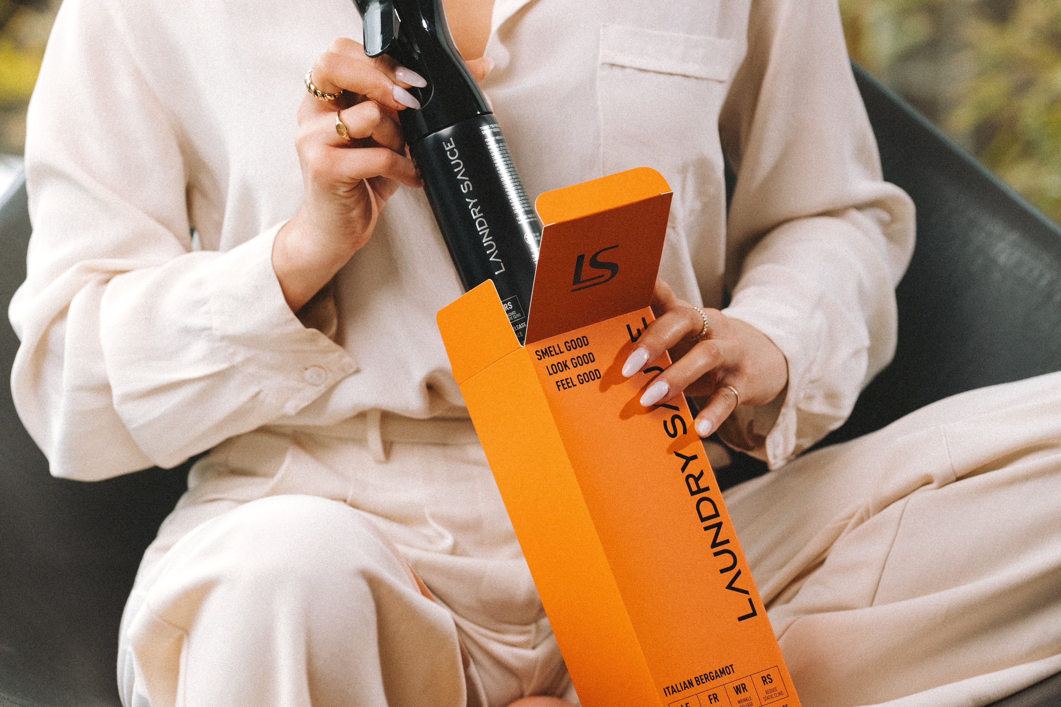 Woman in a cream outfit holding the Laundry Sauce Fabric Refresher Spray and its orange box