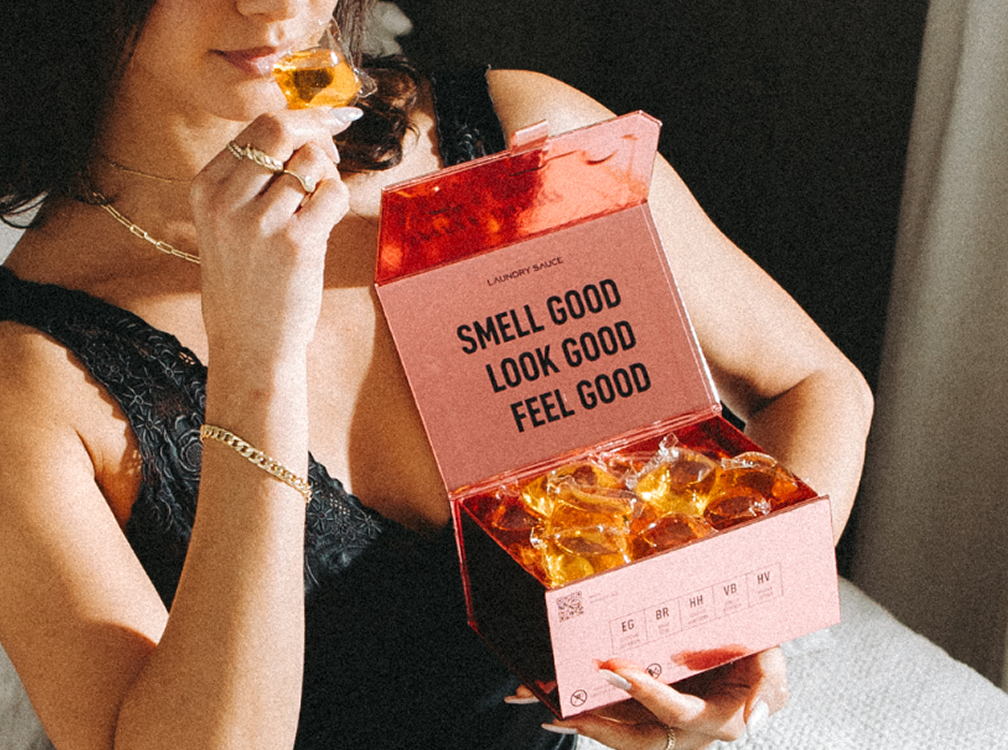 Woman holding an open pink box of Laundry Sauce Laundry Detergent Pods and smelling one