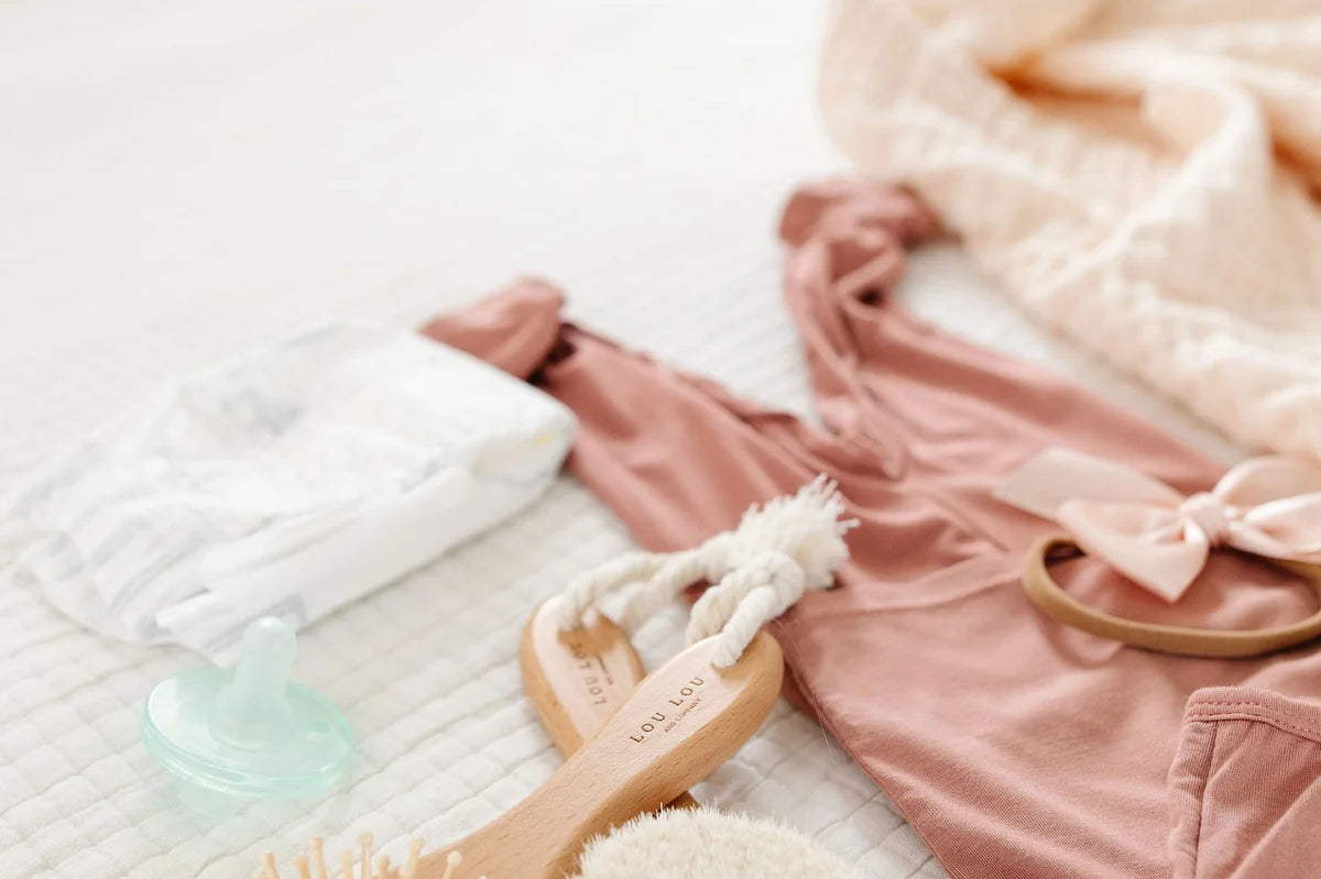 Lou Lou & Company Baby Hair Brush Set, wooden brushes laid on a bed with a mauve baby outfit and pacifier