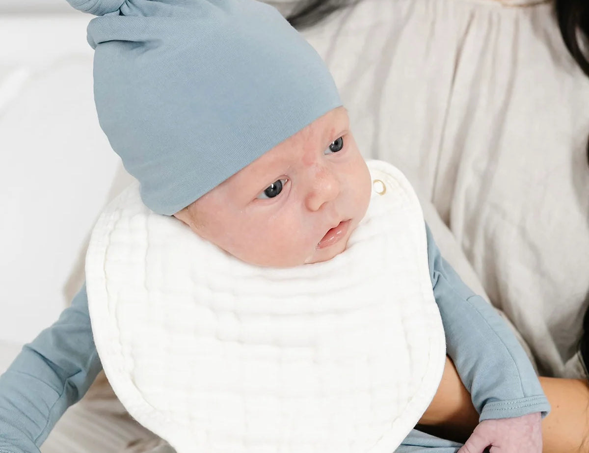 Newborn in a blue knotted hat wearing a white Lou Lou & Company Cloud Muslin Bib
